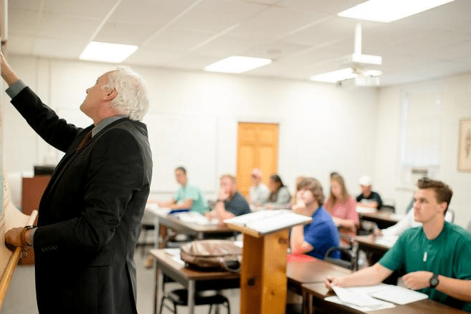 A teacher in a suit points to a map at the front of a classroom, while students sitting at desks listen and take notes. The classroom is bright with white walls and fluorescent lighting.