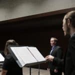 Three adults look down at sheet music while singing