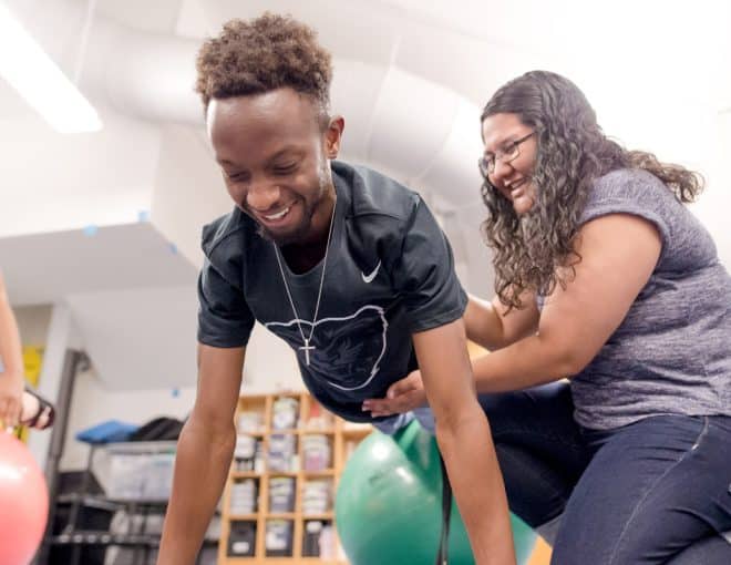 A young man smiles while balancing on a large exercise ball, supported by a woman kneeling beside him. Another person holds a red exercise ball nearby. The scene takes place in a bright, indoor gym or therapy room.