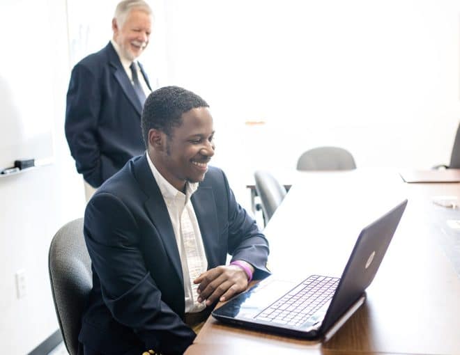 A man in a suit sits at a conference table, smiling at a laptop screen. Another man in a suit stands behind him, also smiling. The room is bright with a whiteboard and framed art on the wall.