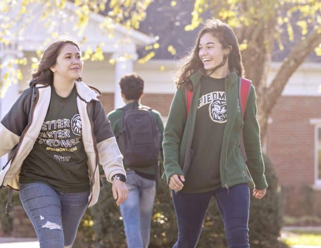 Two young women smile and walk together outside on a sunny day, wearing jeans and T-shirts. A young man with a backpack walks ahead of them towards a brick building with large windows. Trees with fresh leaves are visible.