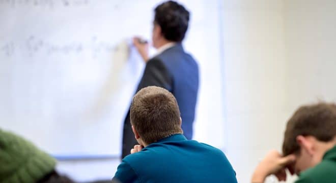 Students sit in a classroom, facing a teacher who is writing on a whiteboard. The teacher is out of focus, while students are in the foreground, paying attention.
