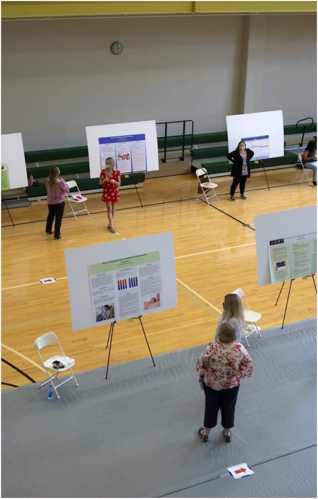People stand near poster boards on easels arranged in a gymnasium for a presentation or science fair. Some individuals are talking while others observe the posters. Folding chairs are placed near each display.