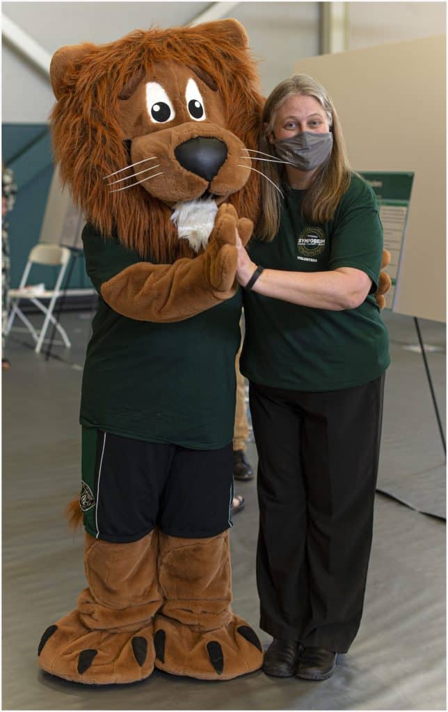 A person wearing a face mask poses and smiles with someone in a friendly lion mascot costume. Both are standing indoors on a gym floor and wearing green shirts.