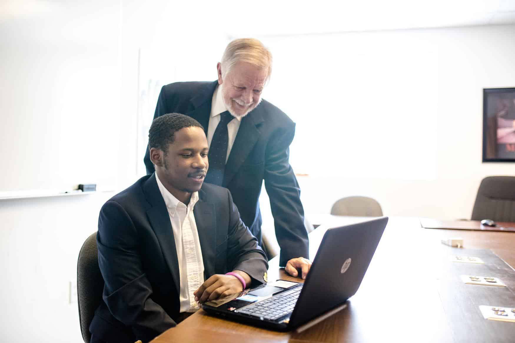 Two men in business suits are in an office. One man is seated and working on a laptop, while the other man stands beside him, smiling and looking at the laptop screen.