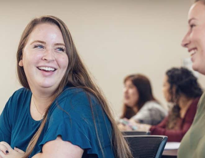 Two young women sit in a classroom, smiling and talking with each other. Other students are seated and engaged in the background, creating a friendly and relaxed learning environment.