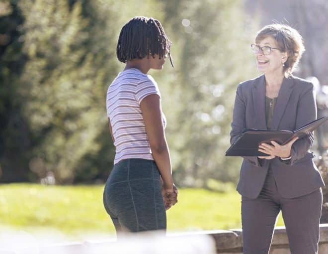Two women stand outdoors talking. One woman is holding a folder and smiling, while the other faces her, listening attentively. The background is sunlit and leafy.