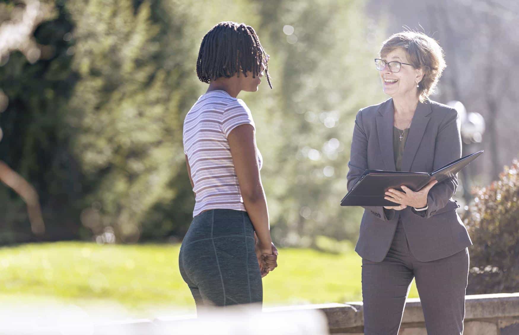 Two women stand outdoors talking. One woman is holding a folder and smiling, while the other faces her, listening attentively. The background is sunlit and leafy.