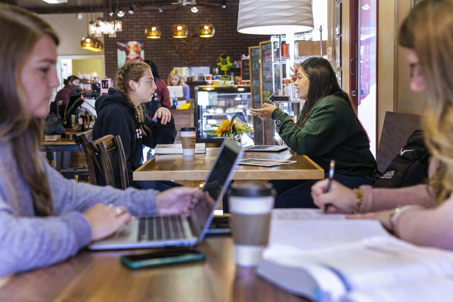 Four young women sit in a cozy café with laptops, notebooks, and coffee cups. Two talk across a table, while two others study and write at the front. Warm lighting and a counter are visible in the background.