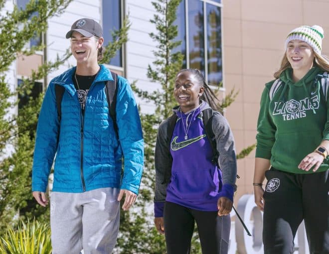 Three students wearing casual clothes and backpacks walk together outside on a sunny day, smiling and talking. There are green trees and a modern building in the background.