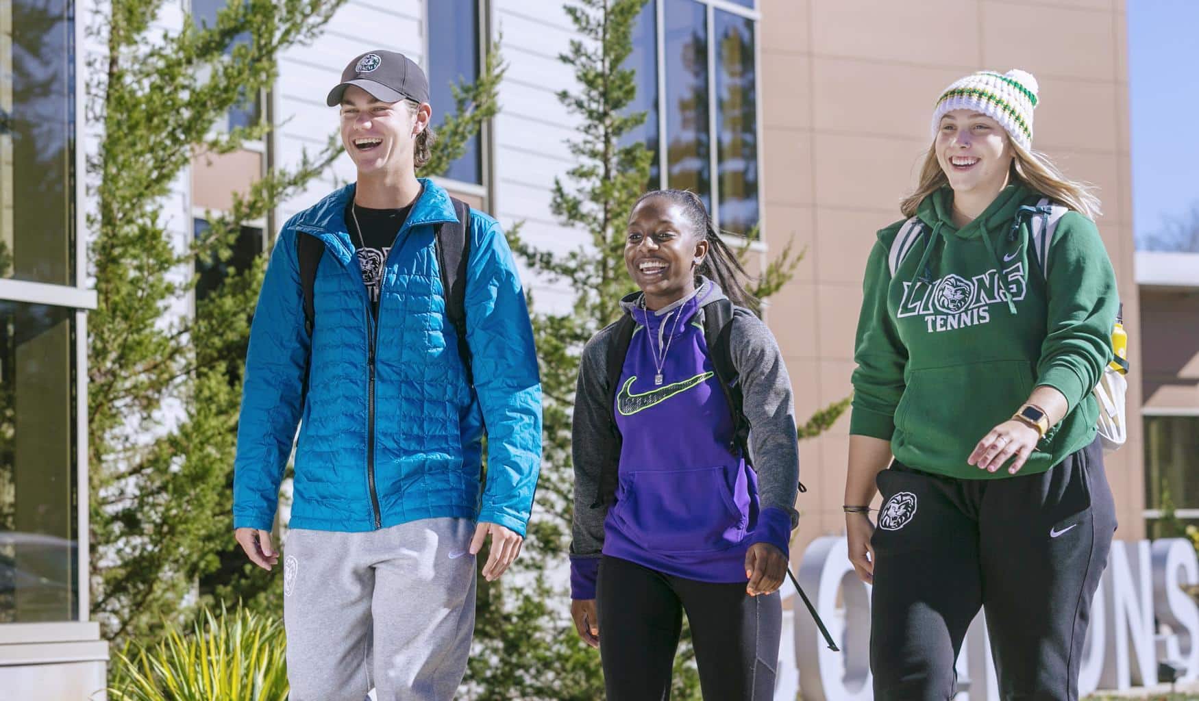Three students wearing casual clothes and backpacks walk together outside on a sunny day, smiling and talking. There are green trees and a modern building in the background.