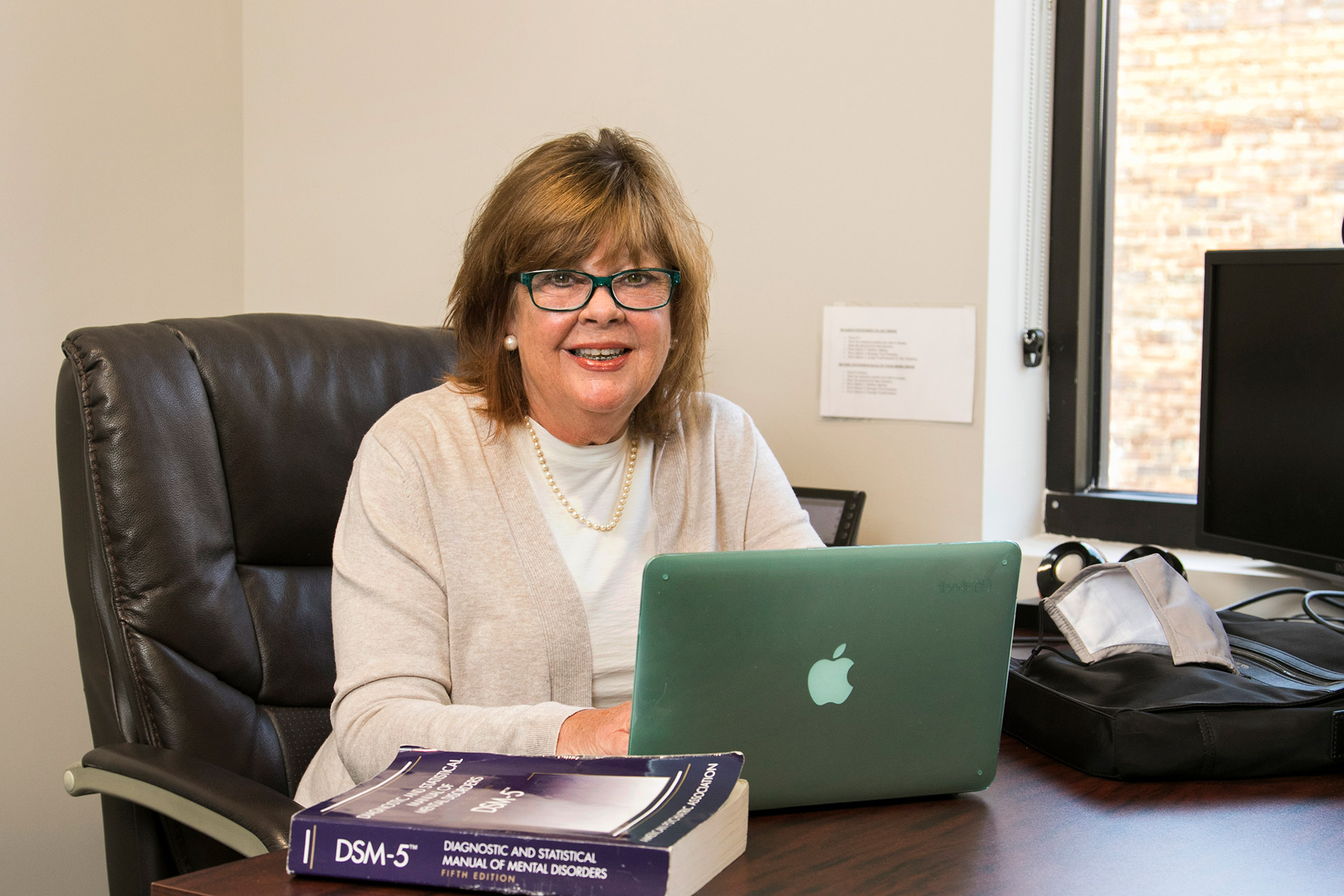 A woman with glasses and shoulder-length hair sits at a desk, smiling at the camera while using a green Apple laptop. A DSM-5 book and a black bag are on the desk in front of her.