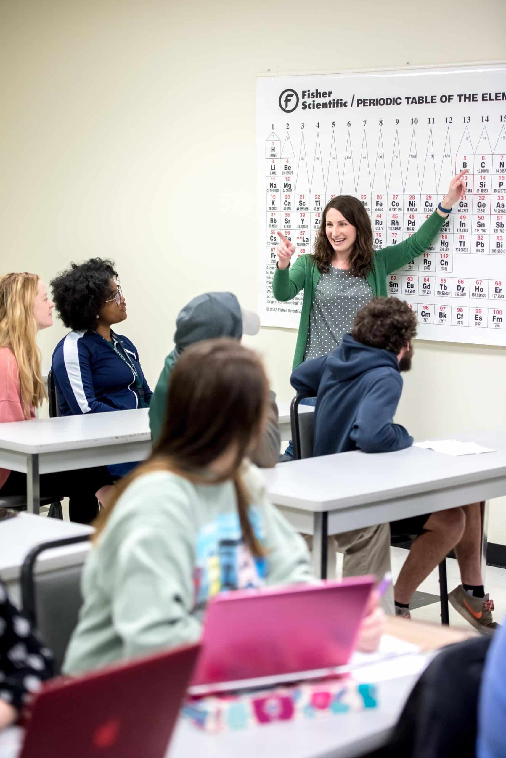 A student points to the periodic table of elements on the wall, while other students watch.