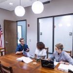 Three students sit at a wooden table in discussion, with laptops and textbooks open.