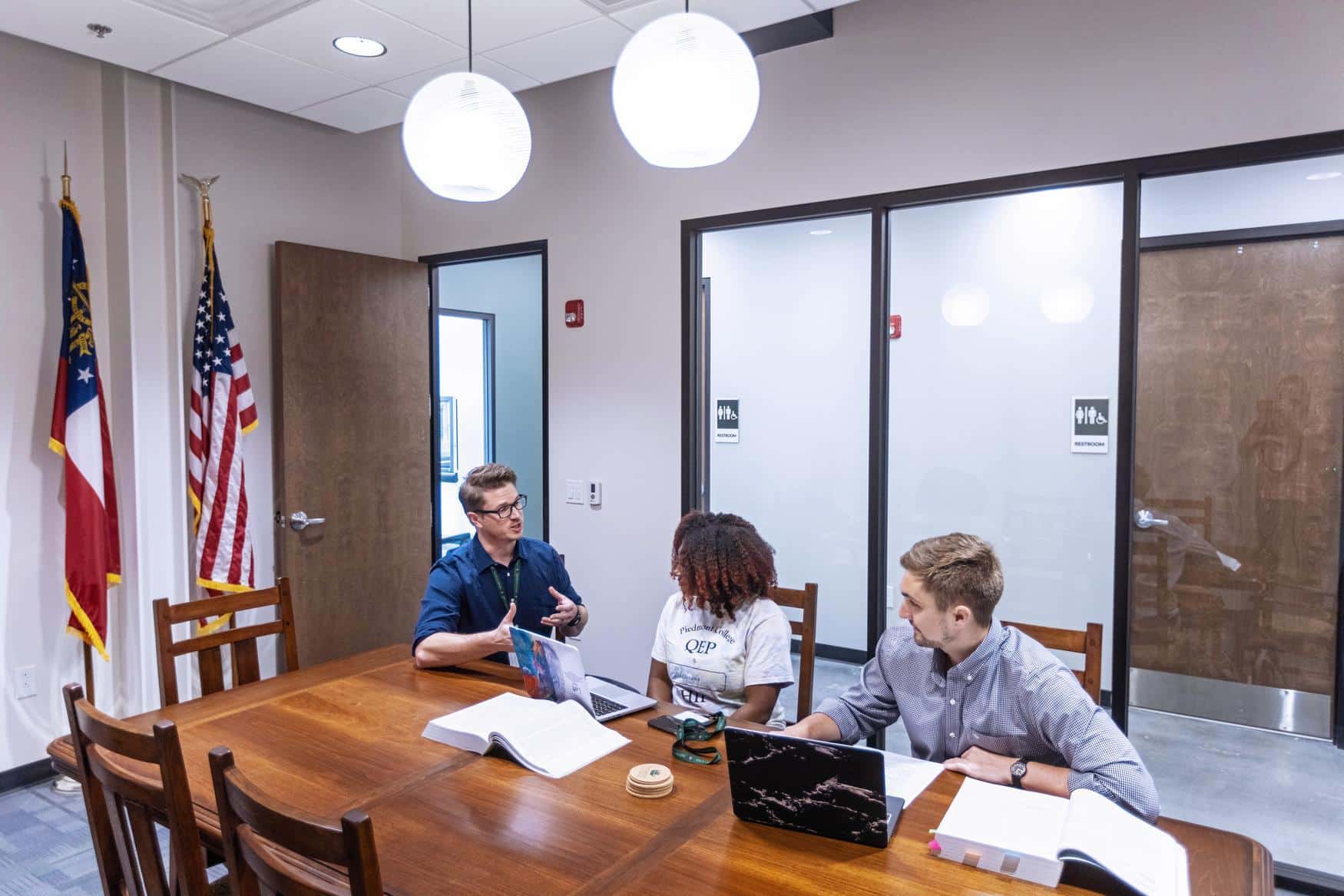 Three students sit at a wooden table in discussion, with laptops and textbooks open.