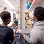Two students interact with and move an educational skeleton model. Other students are visible in the background.