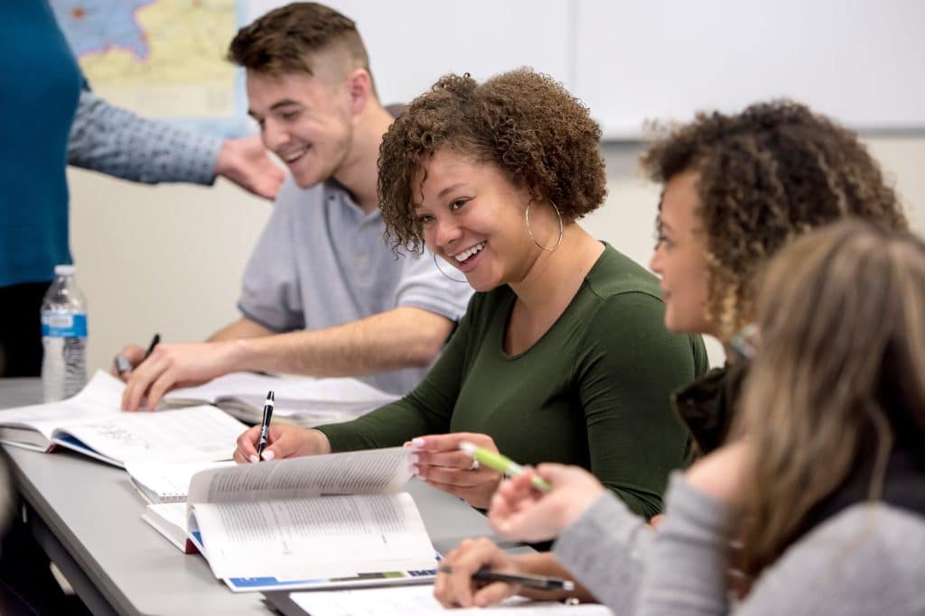 A group of young adults smile and laugh at something off-camera. They are seated at a narrow desk in a classroom.