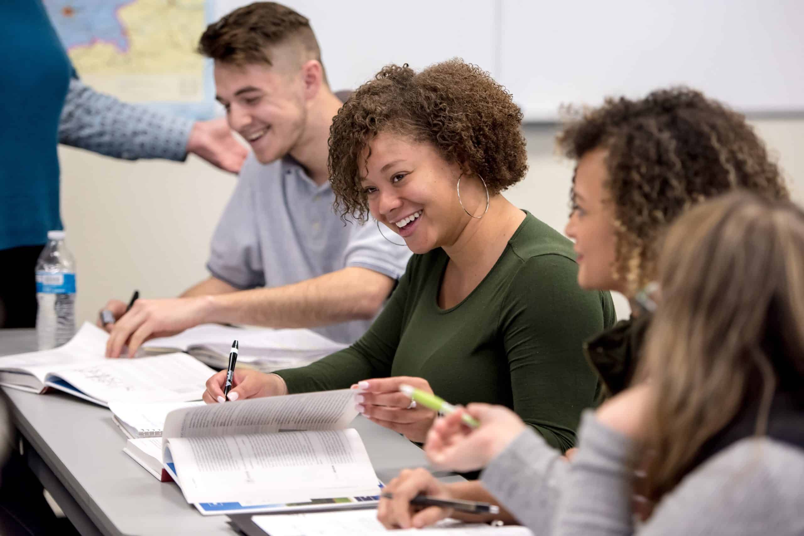 A group of young adults smile and laugh at something off-camera. They are seated at a narrow desk in a classroom.