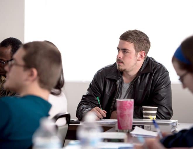 Young man wearing a leather jacket taking notes. Other students are blurred around him, also taking notes.