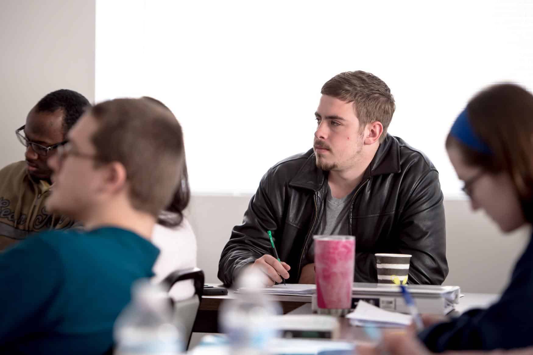 Young man wearing a leather jacket taking notes. Other students are blurred around him, also taking notes.