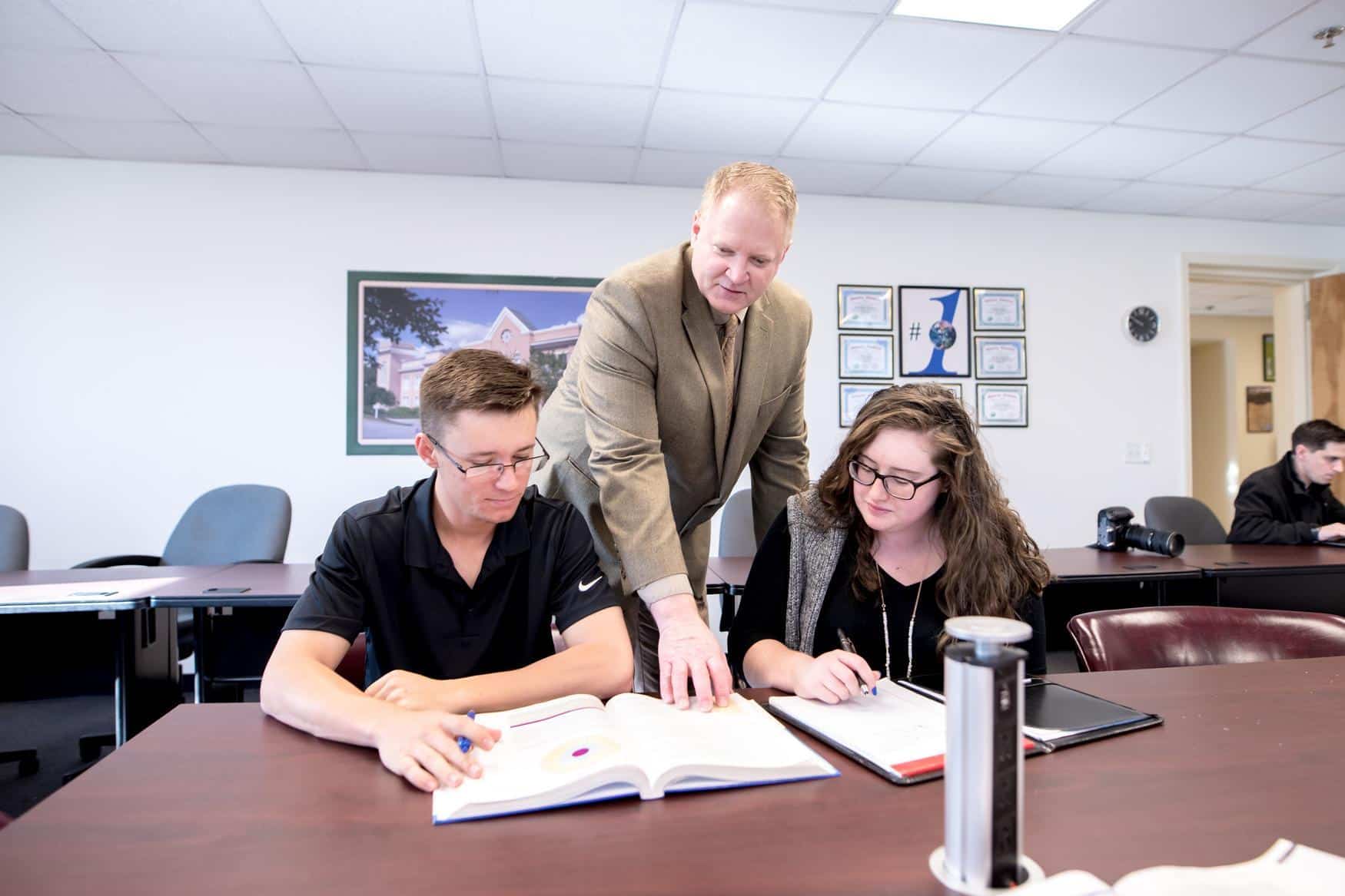 Man in a suit leaning between two students, pointing at an open book