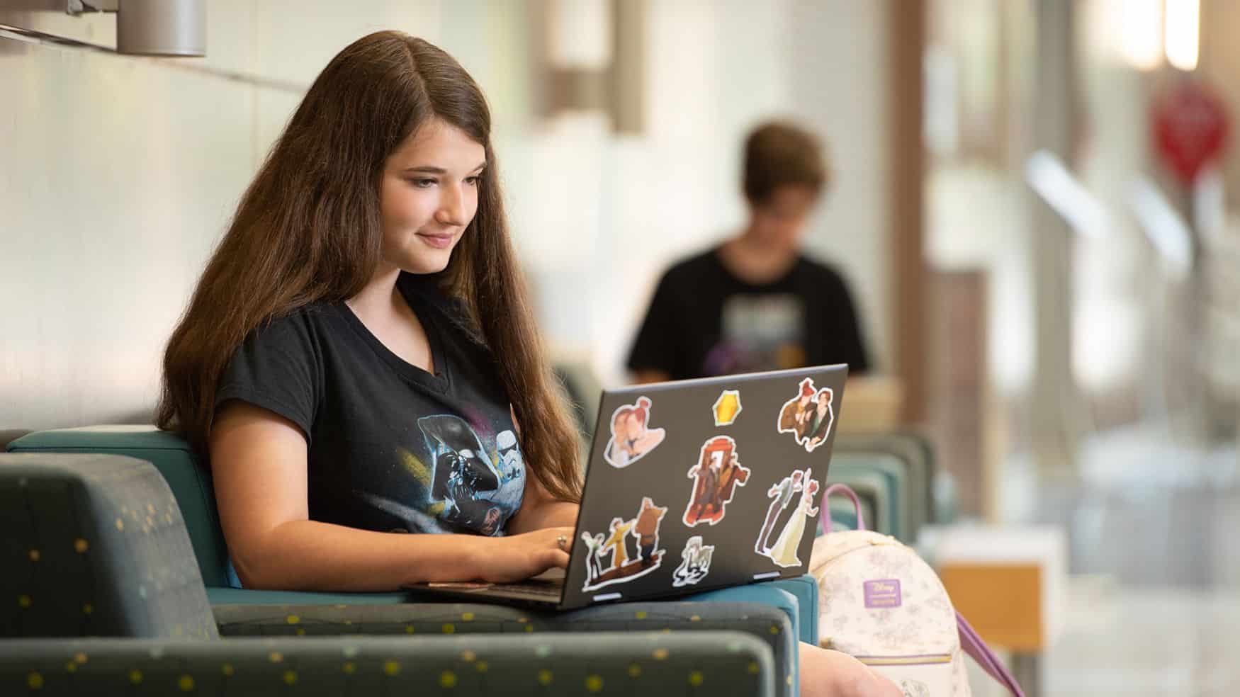 Young woman with long brown hair sitting in a seat with a laptop.