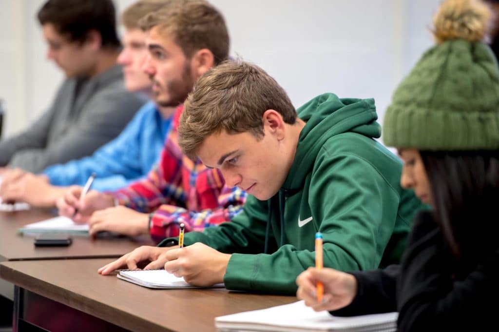 A group of students sits at desks, writing in notebooks. The focus is on a student in a green hoodie, concentrating on coursework for a minor in actuarial science. Other students are in the background, also taking notes.