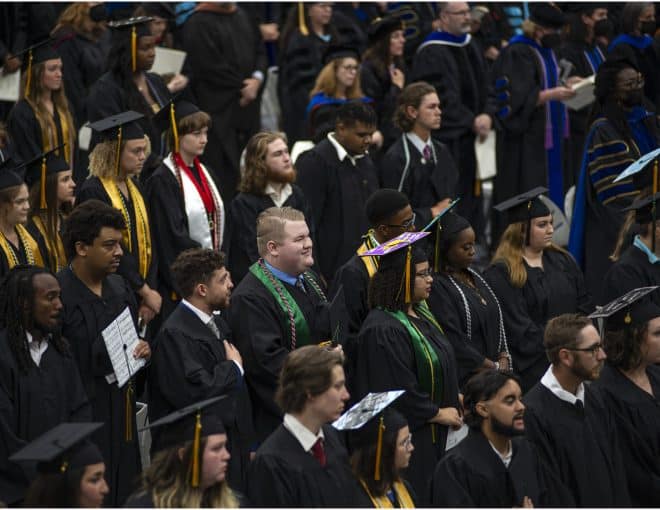A large group of graduates in caps and gowns stand during a graduation ceremony. Some wear honor cords or decorated caps, and faculty members in academic regalia are visible in the background.