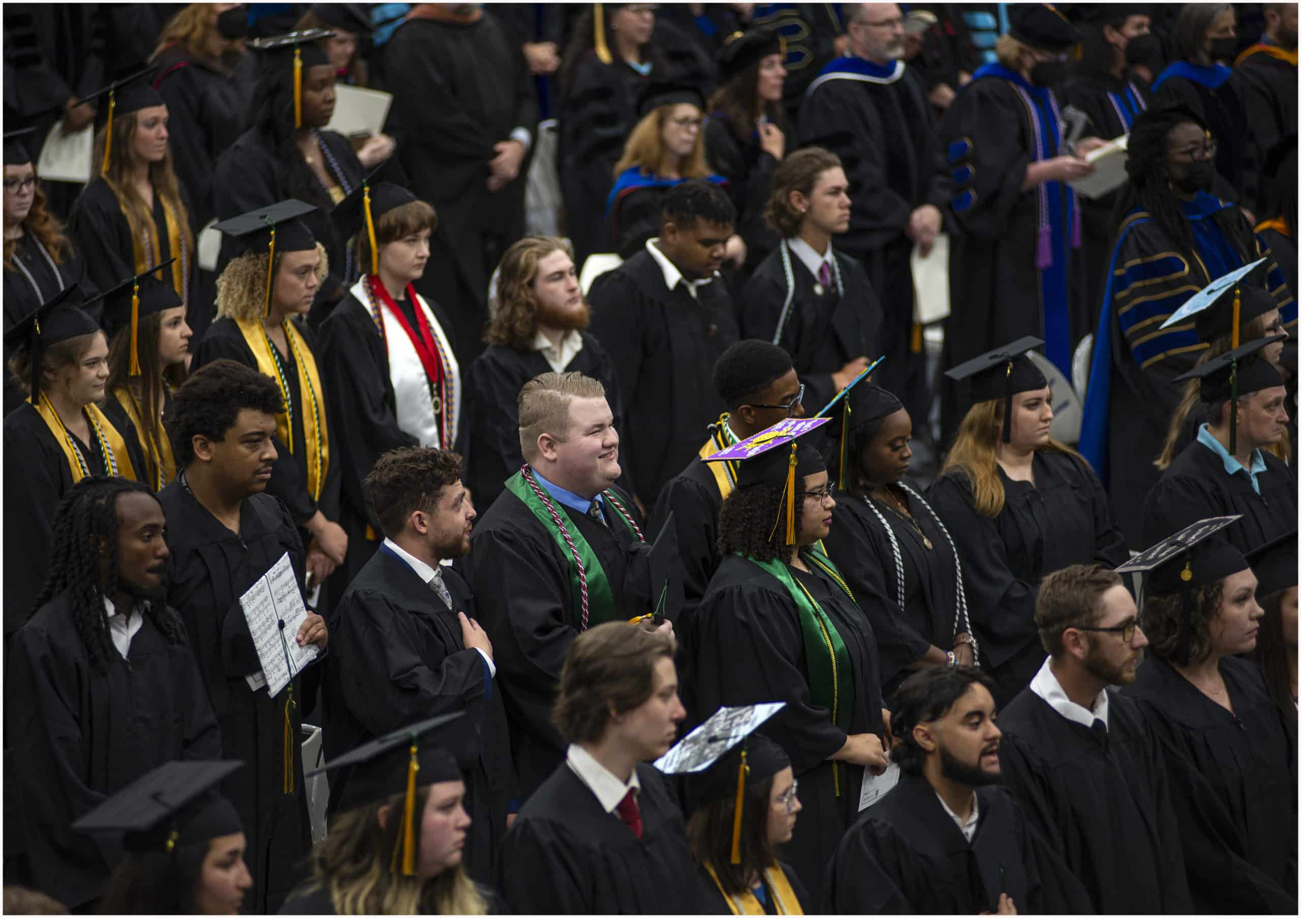 A large group of graduates in caps and gowns stand during a graduation ceremony. Some wear honor cords or decorated caps, and faculty members in academic regalia are visible in the background.