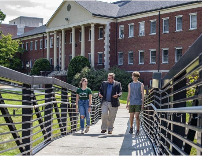 Three people—a man in a blazer and two young adults—walk and talk on a bridge leading to a large brick building with white columns on a sunny day.