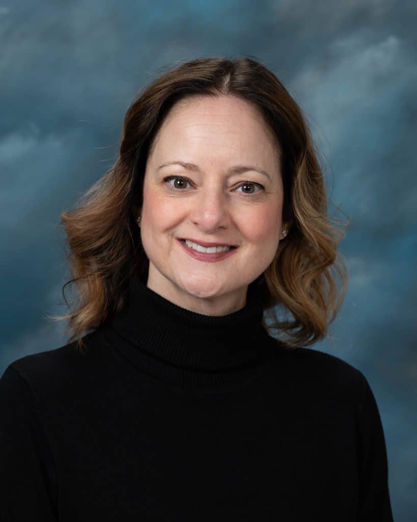 A woman with shoulder-length brown hair and light skin smiles at the camera. She is wearing a black turtleneck sweater, with a blue and gray cloudy background behind her.