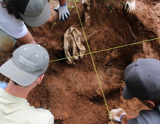 Three people in hats and gloves are digging and examining a dirt excavation site, marked with yellow string in a grid. One person wears a “Piedmont University” hat. A cloth is partially buried in the center.