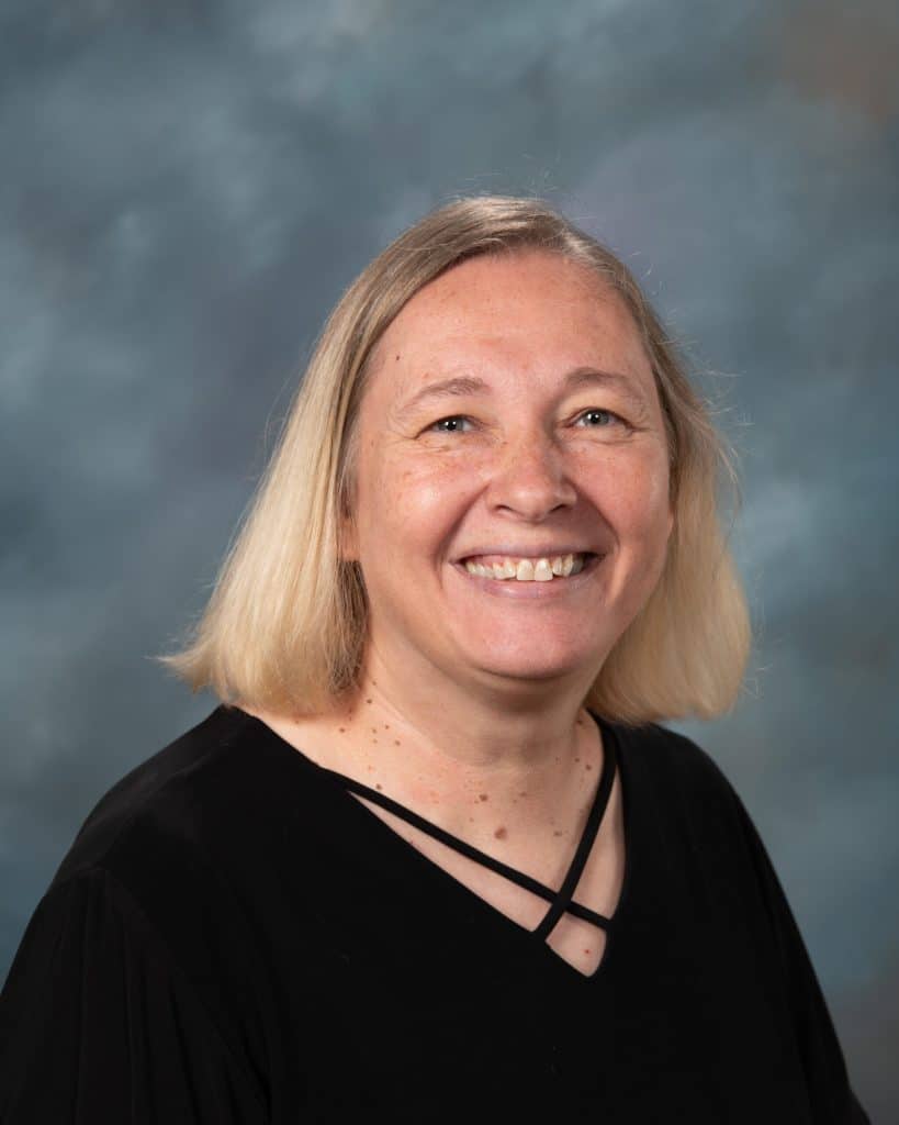 A woman with straight, shoulder-length blonde hair and light skin is smiling at the camera. She is wearing a black top with crisscross straps, in front of a blue-gray mottled background.