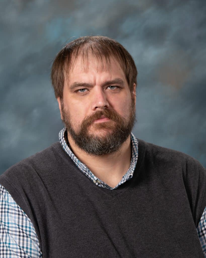 A man with a beard and mustache wearing a gray sweater over a blue and white checkered shirt, sitting in front of a mottled gray-blue background. He has a serious expression.