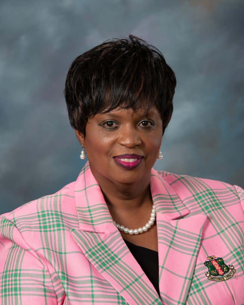 A woman with short dark hair wears a pink plaid blazer with green accents, a black top, a pearl necklace, and pearl earrings, smiling in front of a blue-gray background.