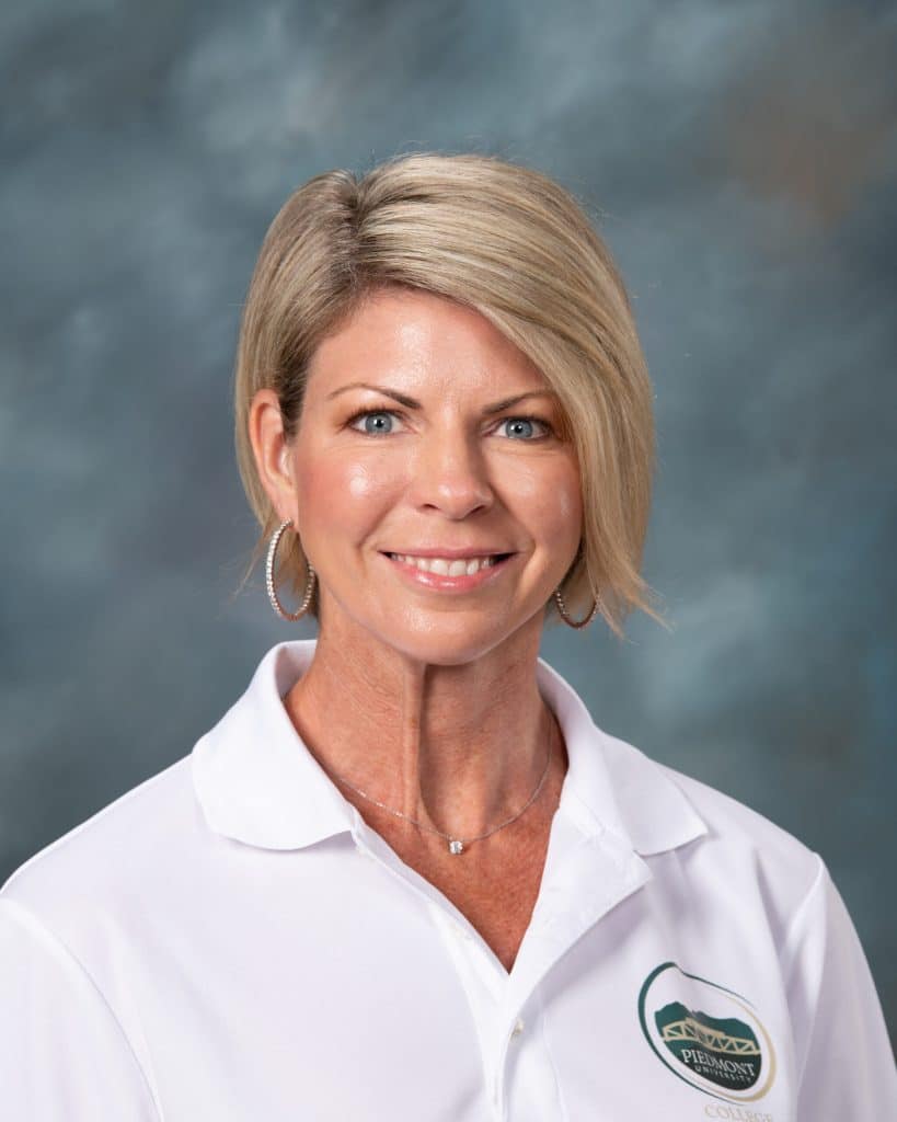 A woman with short blonde hair, wearing hoop earrings and a white collared shirt with a green logo, smiles at the camera against a blue-gray background.