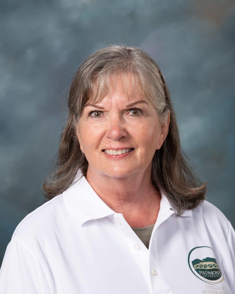 A woman with gray hair and bangs smiles at the camera, wearing a white collared shirt with a Piedmont College logo, against a blue-gray mottled background.
