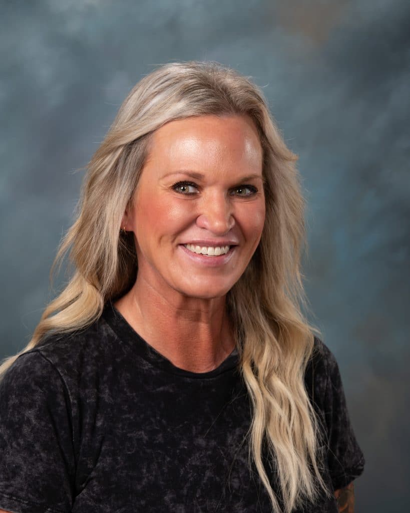 A smiling woman with long, wavy blonde hair wearing a black, short-sleeve top, posing in front of a blue and gray mottled background.