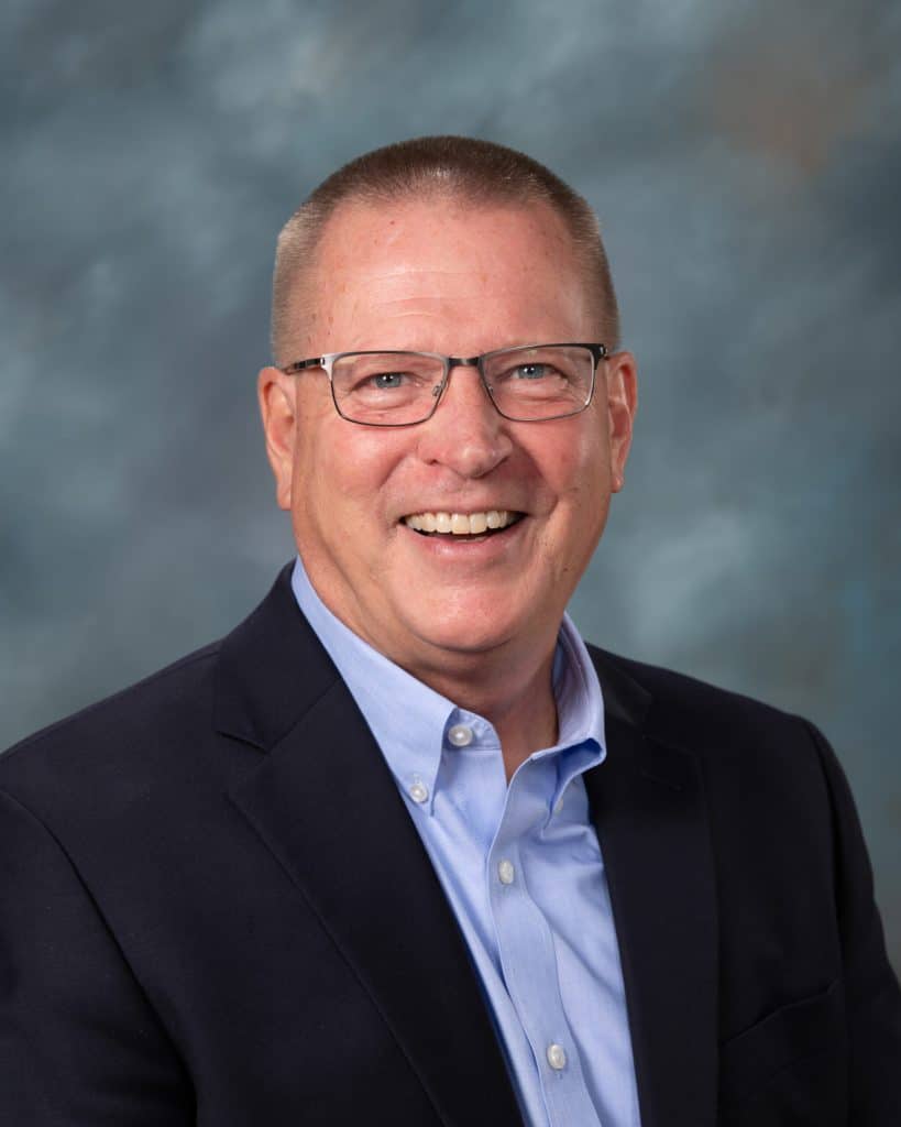 A middle-aged man with short hair, glasses, and a blue dress shirt under a dark suit jacket, smiling at the camera in front of a softly blurred blue-gray background.