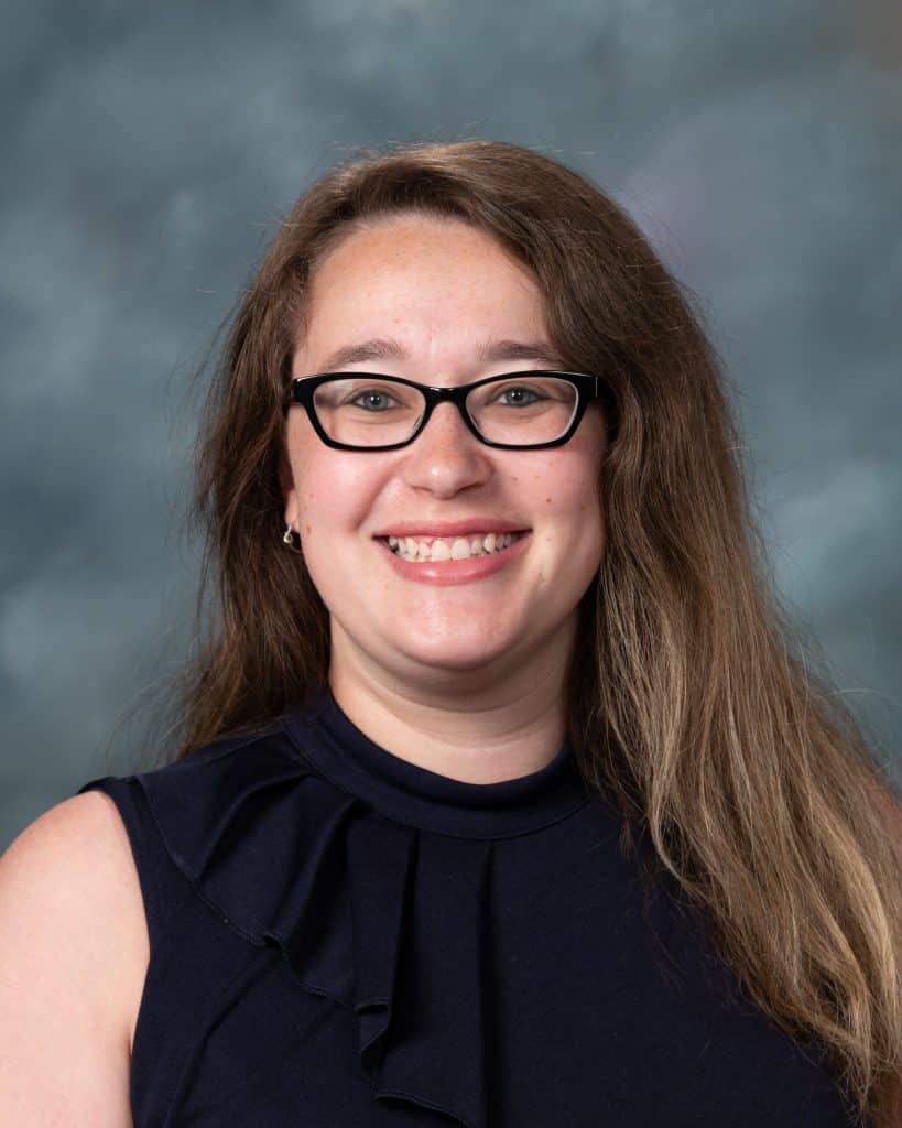 A woman with long light brown hair and glasses smiles at the camera. She is wearing a sleeveless navy blouse. The background is a soft, blurred gray and blue gradient.