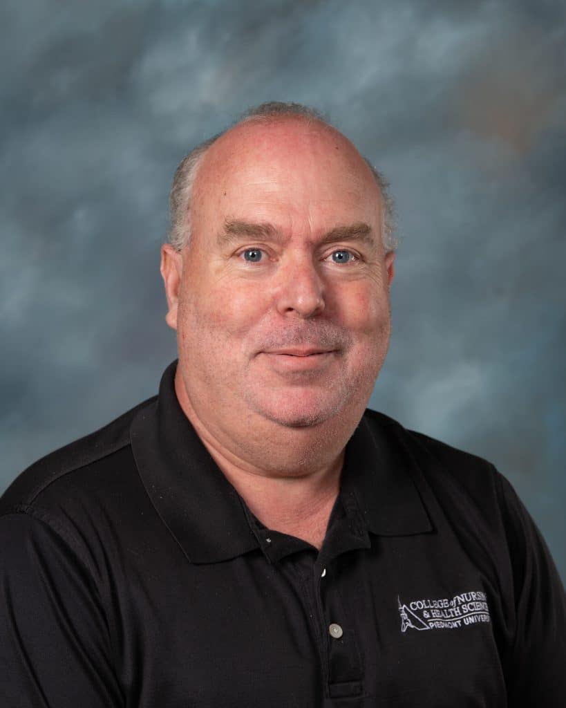 A middle-aged man with light skin, short thinning gray hair, and blue eyes, wearing a black polo shirt, smiles at the camera against a cloudy gray-blue backdrop.