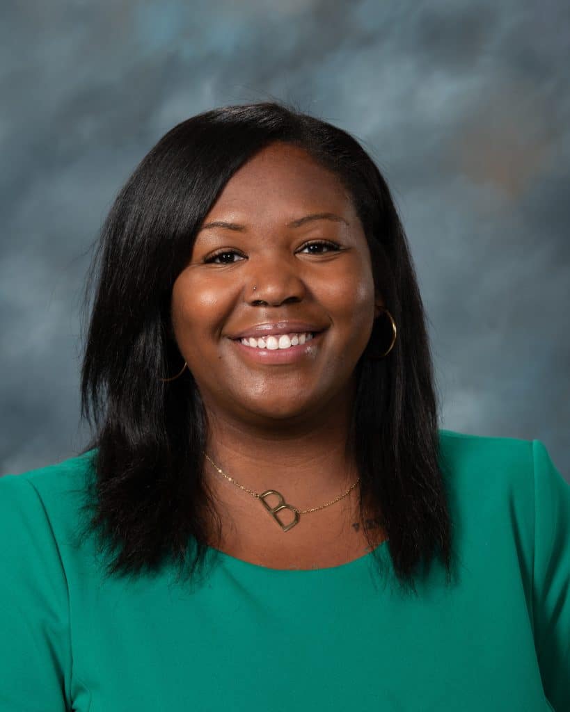 A woman with straight black hair, wearing a green top, gold hoop earrings, and a gold necklace, smiles at the camera against a softly blurred gray and blue background.