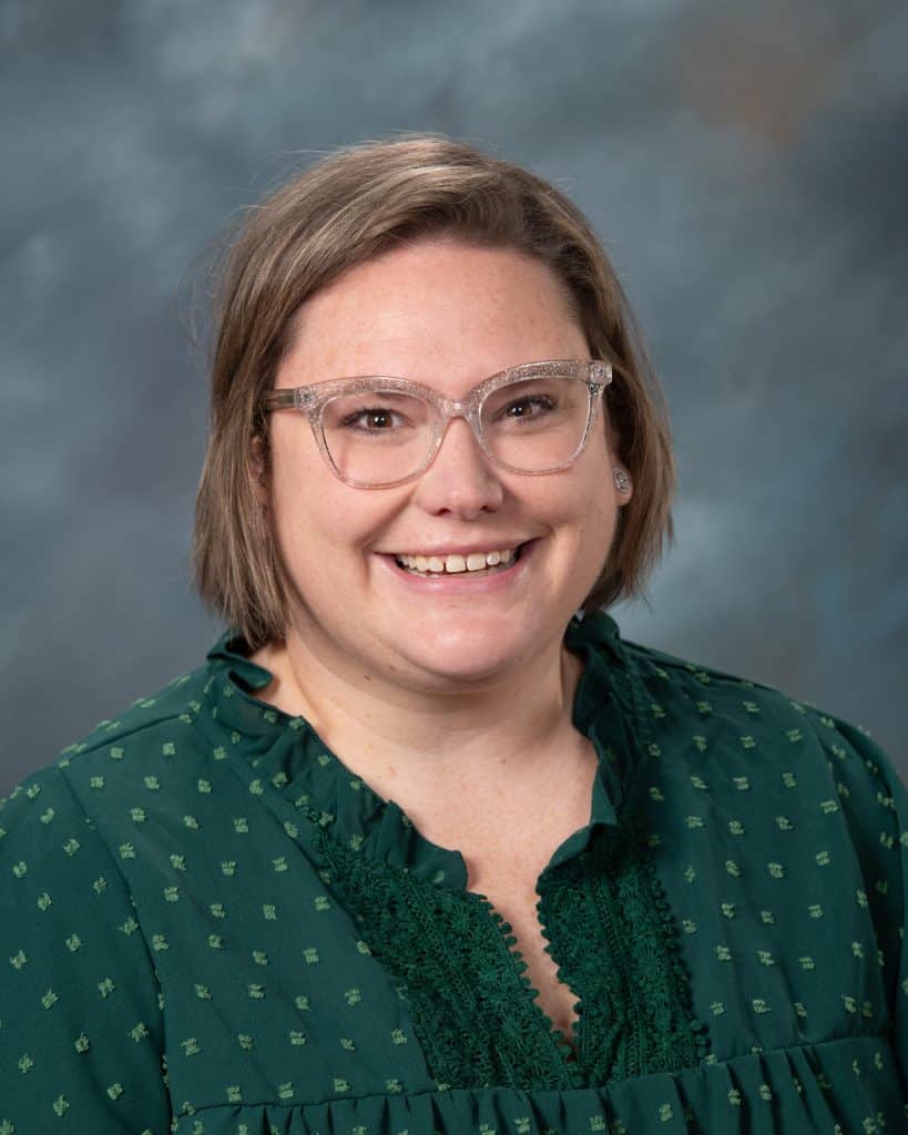 A woman with short light brown hair and clear glasses smiles at the camera. She is wearing a dark green blouse with a textured pattern. The background is a soft, blurred mix of blue and gray tones.