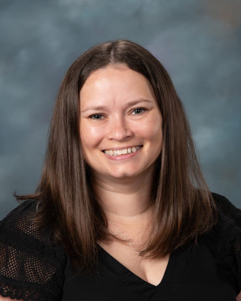 A woman with straight brown hair and a black top smiles at the camera in front of a cloudy blue-gray background.