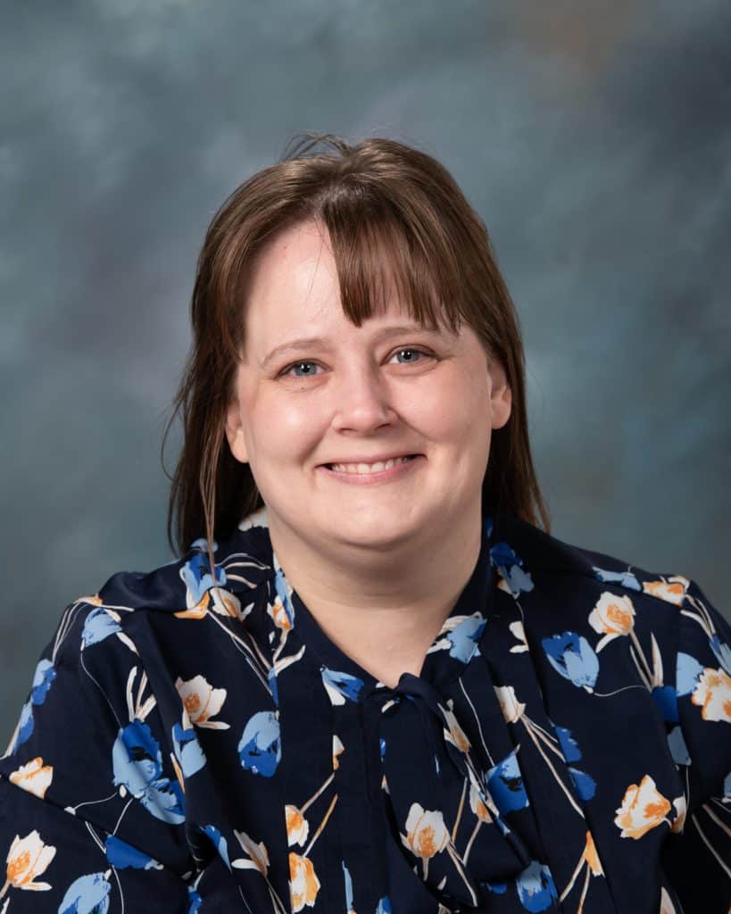 A woman with straight brown hair and bangs smiles at the camera. She is wearing a dark blouse with a blue and orange floral pattern, and the background is a soft, blurred blue-gray.