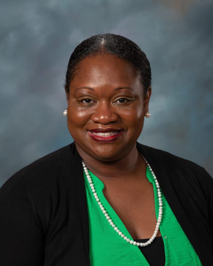 A woman with short, dark hair pulled back, wearing a green top, black cardigan, pearl necklace, and pearl earrings, smiling in front of a blue-gray background.