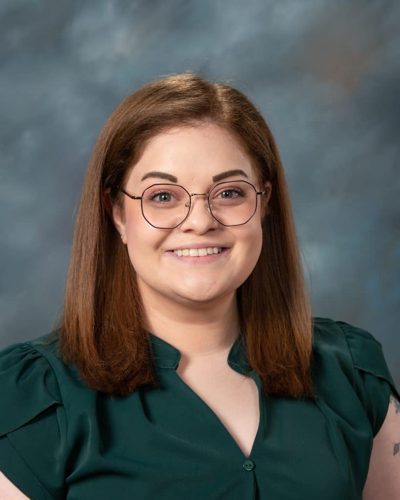 A woman with straight, shoulder-length brown hair and glasses smiles at the camera. She is wearing a dark green blouse and is posed in front of a gray, softly blurred background.