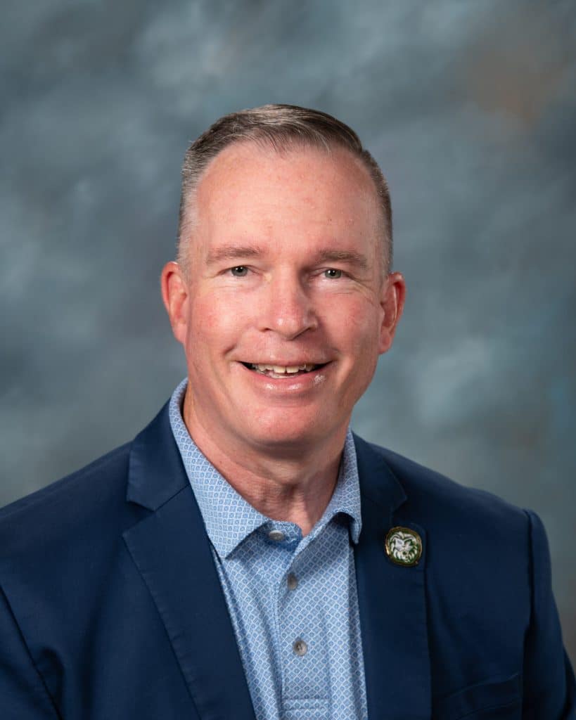 A middle-aged man with short, light hair is smiling. He is wearing a blue blazer, a light blue patterned shirt, and a round pin on his lapel. The background is a blurred mix of gray and blue tones.