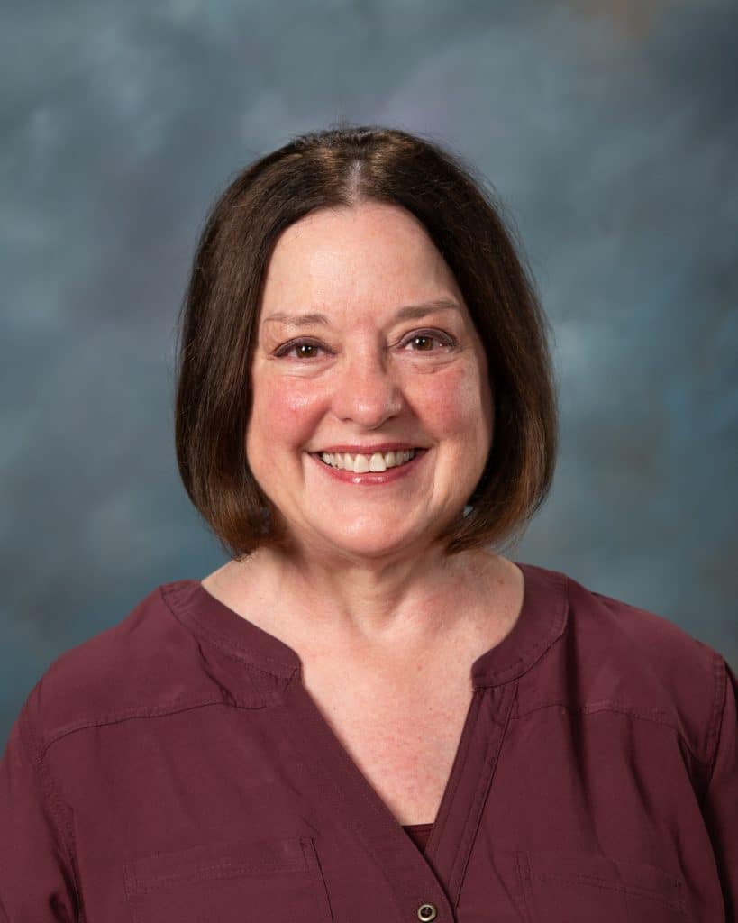 A middle-aged woman with straight brown hair and a maroon shirt smiles at the camera against a blurred blue and gray background.
