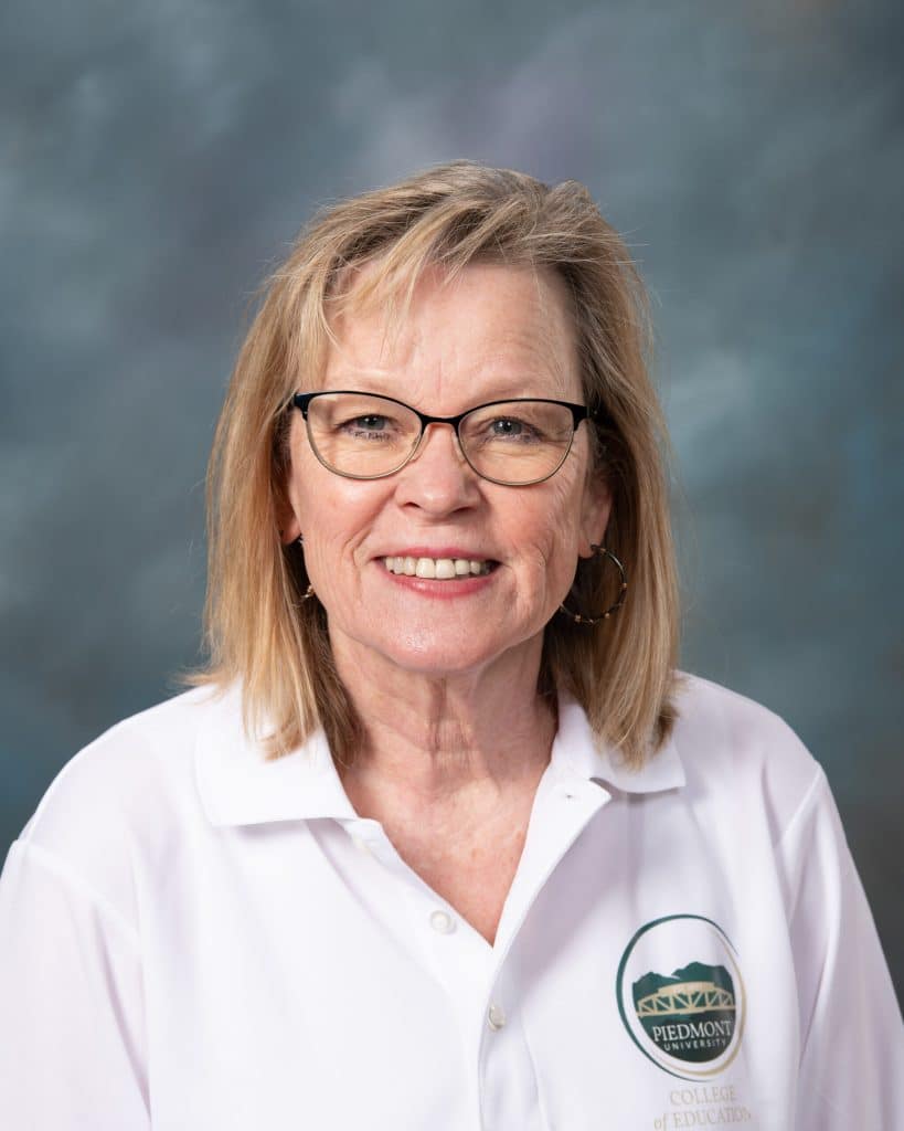 Smiling older woman with blonde hair, glasses, and hoop earrings, wearing a white polo shirt with a Piedmont College of Education logo, posing in front of a blue-gray studio background.